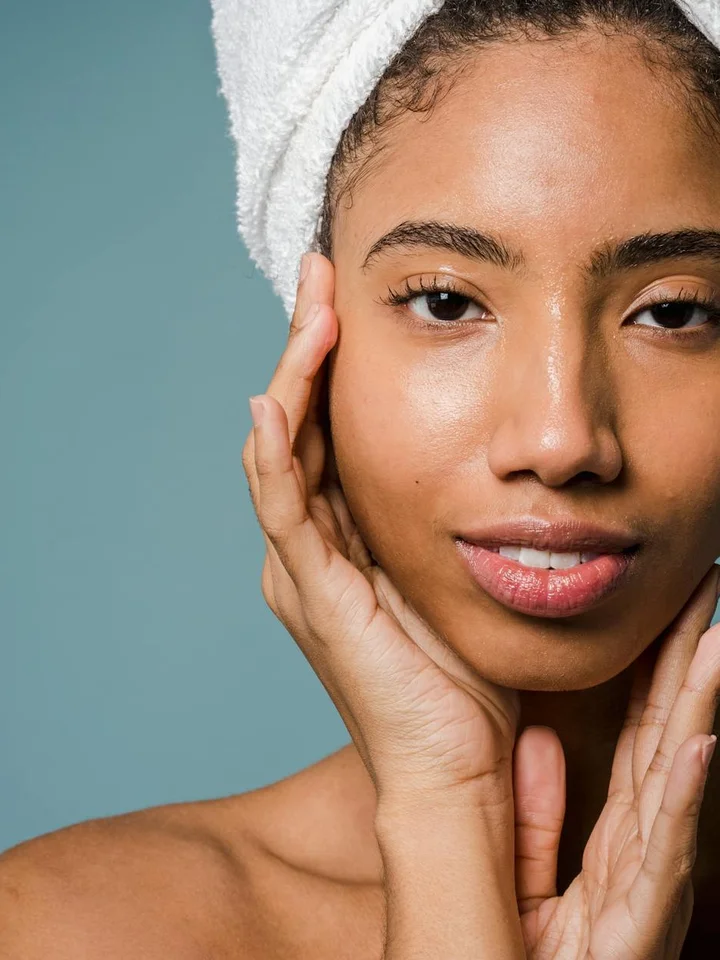 Close-up portrait of a woman with a towel wrapped around her head, touching her face gently.