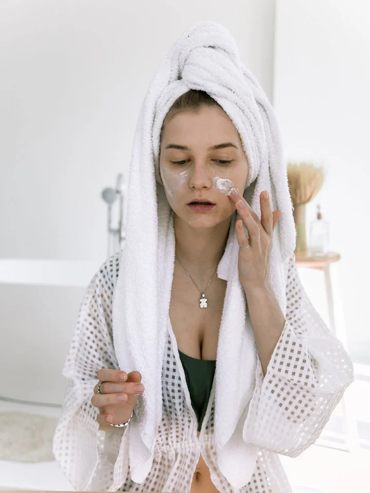 Woman with a towel wrapped around her head applying skincare cream to her face in a bright bathroom