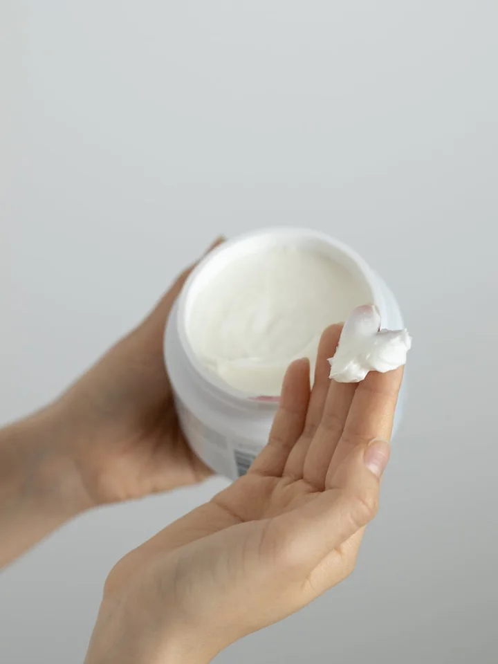 Hands holding an open jar of topical cream with a dab of product on a fingertip