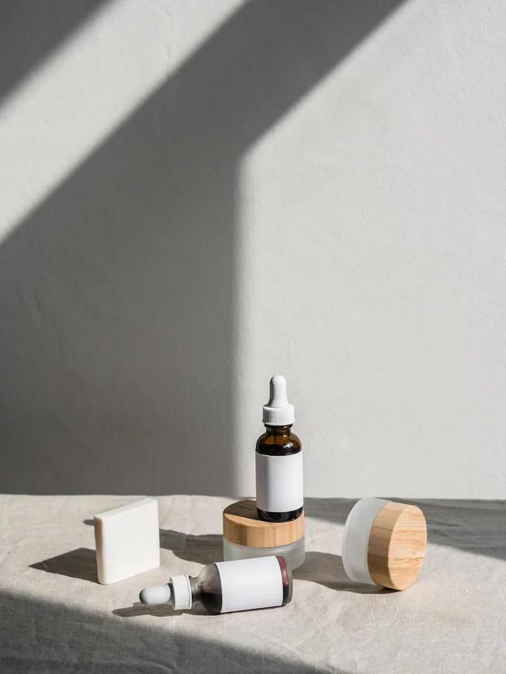 A skincare setup with dropper bottles, a small white bar, and a round container on a light, textured surface with soft shadows.
