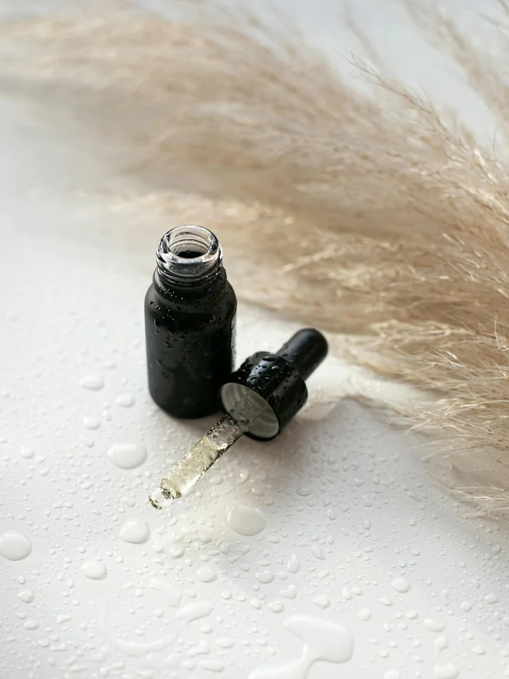 Open dropper bottle of alpha arbutin serum on a white surface with droplets and soft pampas grass in the background.