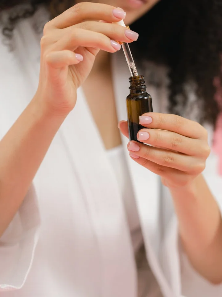 A person holds a small amber dropper bottle and uses a dropper to apply serum to their face.