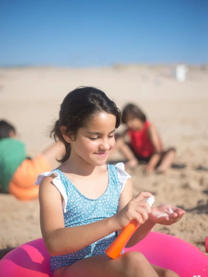 A young girl sits on a pink inflatable ring at the beach, applying sunscreen from an orange bottle to her hand while other children play in the blurred background.