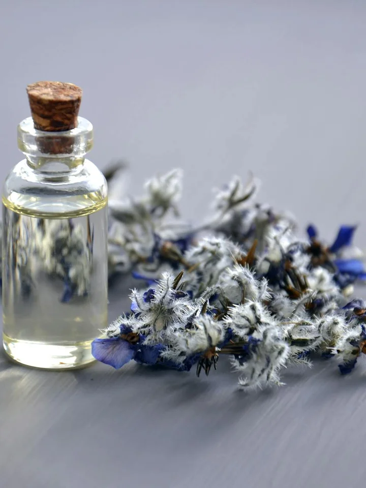 Small glass bottle with a cork stopper placed on a gray surface beside dried blue and white flowers.