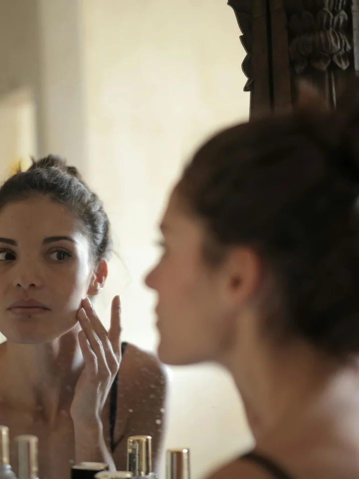 Woman looking at her reflection in a bathroom mirror while touching her cheek.