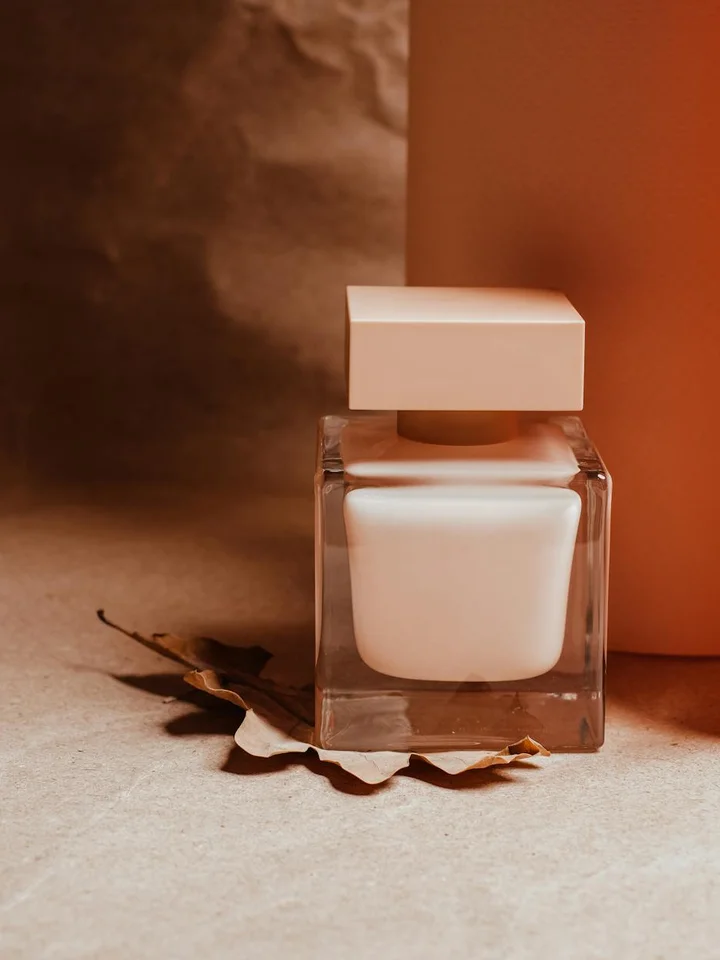 A minimalist square glass bottle with a white cap sits on a warm beige surface with a dried leaf nearby, suggesting clean skincare.