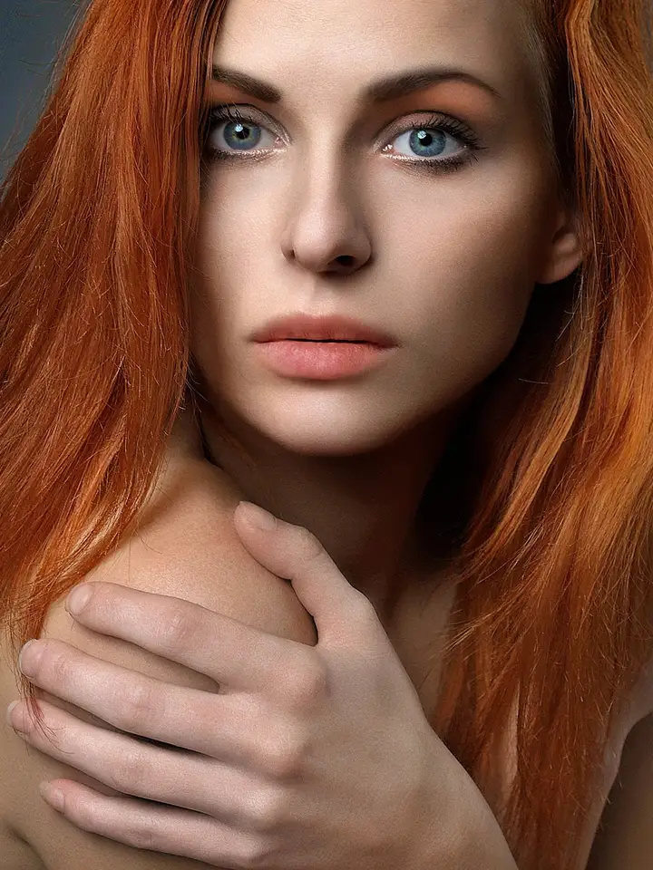 Close-up portrait of a woman with long red hair and blue eyes, looking directly at the camera.