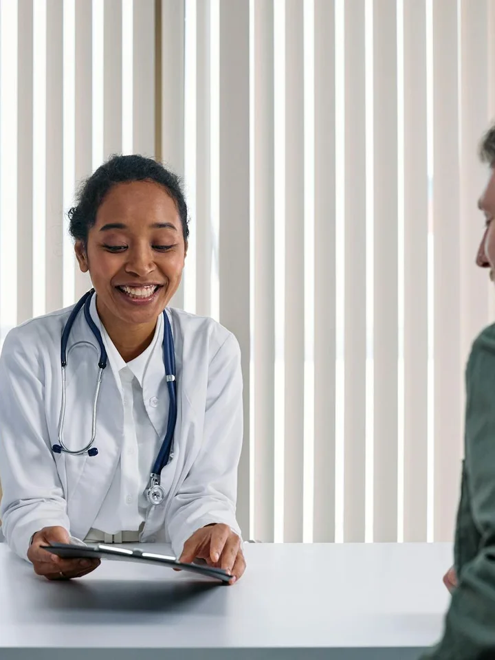 Dermatologist seated at a clinic desk, smiling while showing information on a tablet to a patient.
