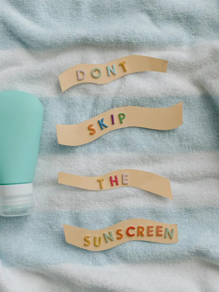 A sunscreen bottle sits on a light blue towel with four beige paper strips, each with colorful letters spelling 'DONT SKIP THE SUNSCREEN'.
