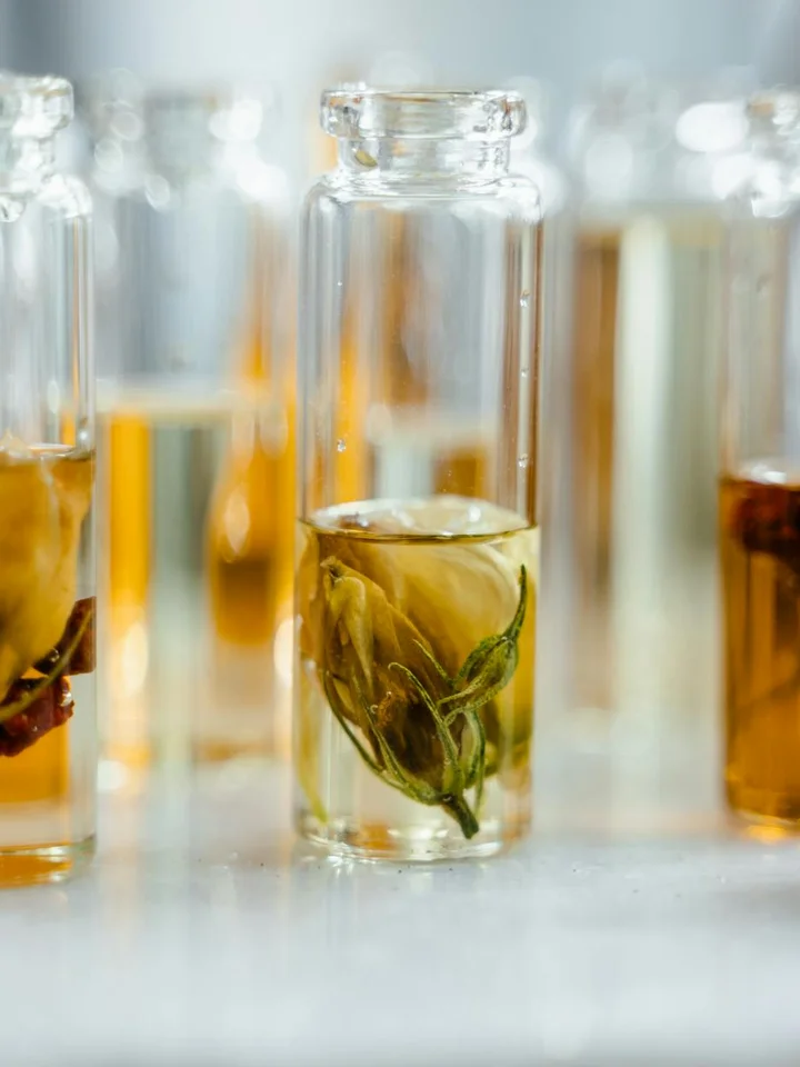 Close-up of small glass bottles containing plant-infused oils and herbs on a bright, white surface