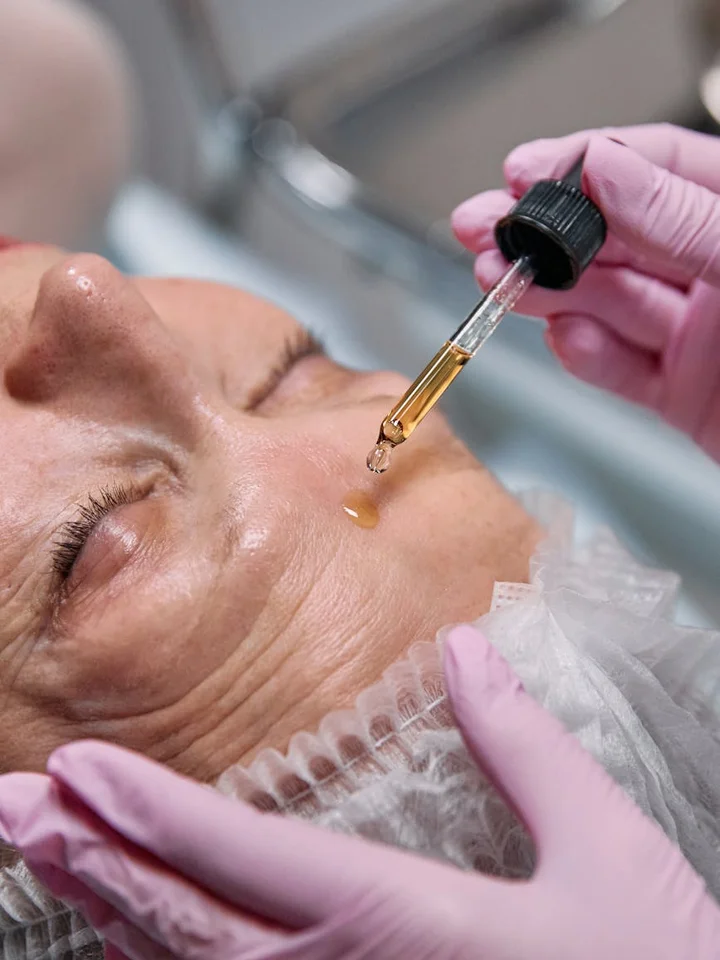 Close-up of a skincare professional wearing pink gloves applying a dropper of serum onto a woman's face during a facial treatment.