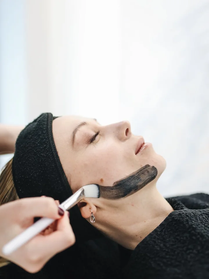 Client lying with eyes closed and wearing a headband while a practitioner applies a dark facial mask with a brush to the cheek.