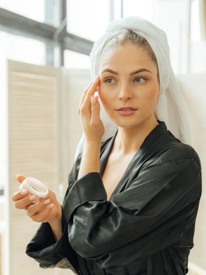 A woman with a towel wrapped around her head applies skincare cream in a bright bathroom.