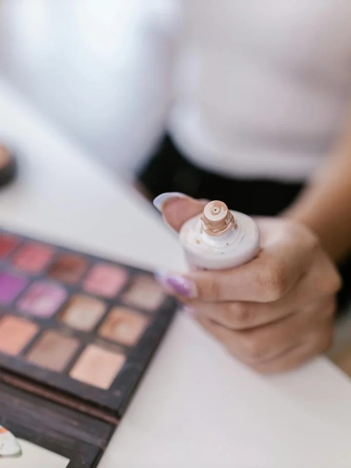 Close-up of hands holding a foundation bottle with a pump, with a makeup palette blurred in the background.