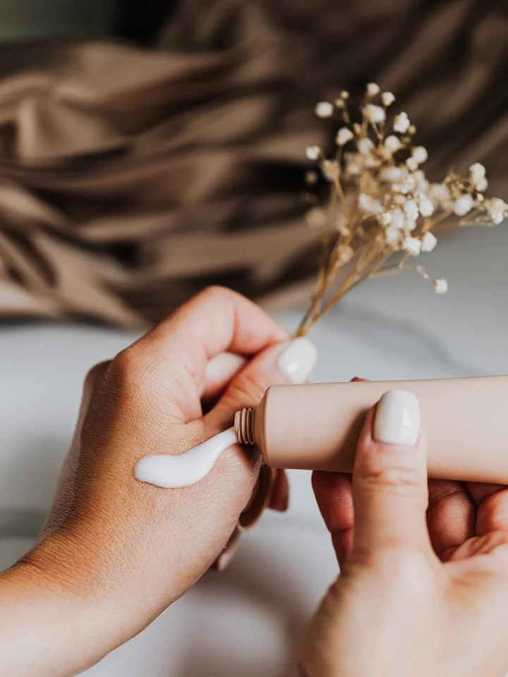 Close-up of hands squeezing a small amount of cream from a tube onto the back of the other hand, with a dried flower in the blurred background.