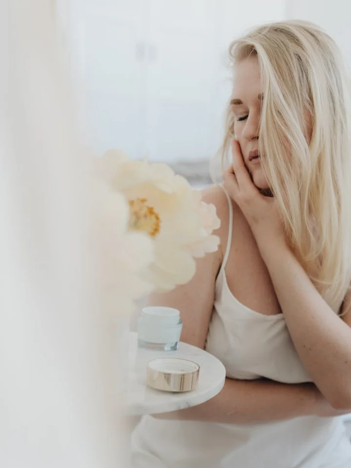 Blonde woman resting her chin on her hand near skincare products on a small table, contemplating a skincare routine.