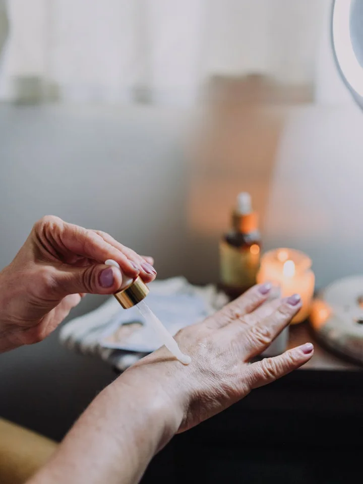 Close-up of a hand receiving a drop of serum from a dropper, with skincare bottles and candles blurred in the background.