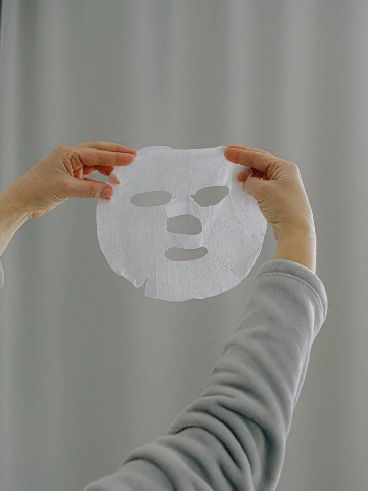 A pair of hands holds up a white facial sheet mask with cut-outs for the eyes, nose, and mouth against a soft gray background.