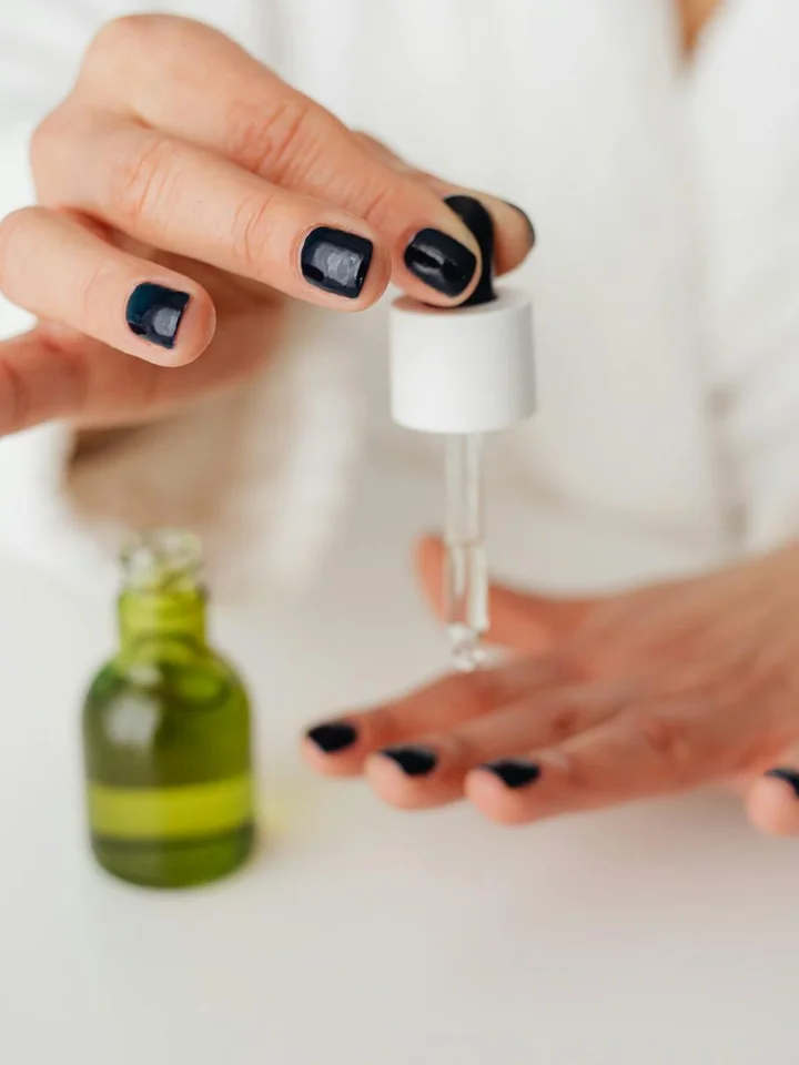 Close-up of a hand with dark nail polish holding a dropper above a small bottle, preparing to apply skincare product.