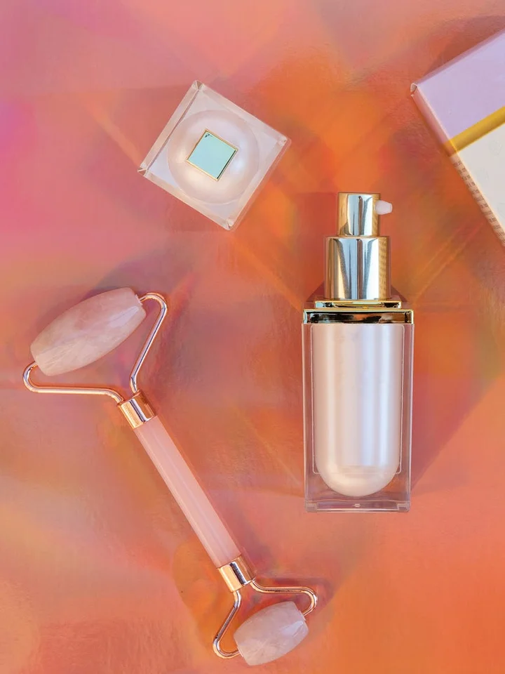 Close-up of skincare tools on a pink-orange background: a glass bottle with pump, a facial roller, a small jar, and a notebook.