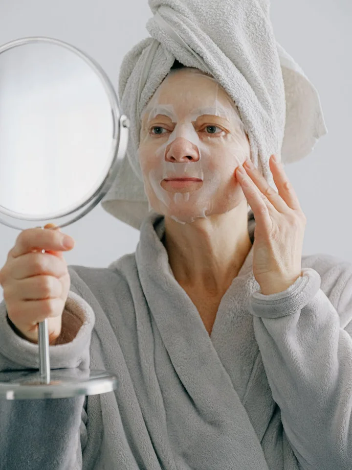 Woman in a facial mask with a towel wrapped around her head, looking into a round mirror while applying skincare.