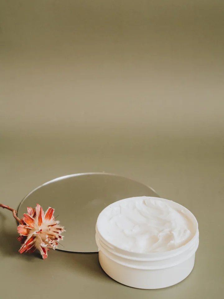 A white jar of cream on a muted green surface with a small dried flower beside it.