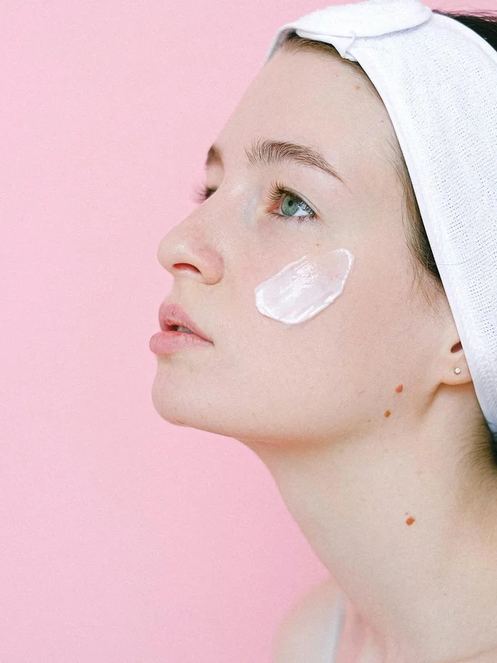 Close-up profile of a young woman with a dollop of tretinoin cream on her cheek, wearing a white headband against a pink background.