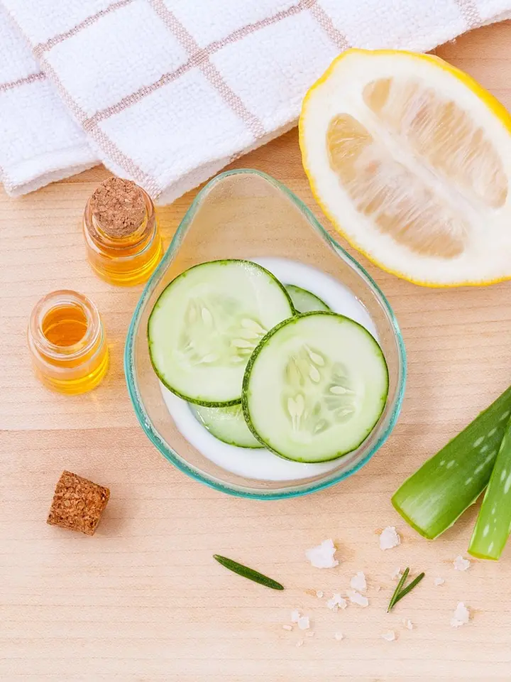 A skincare setup featuring cucumber slices in a glass bowl, a halved lemon, small bottles of oil, a cork, a towel, and aloe-like leaves on a wooden surface