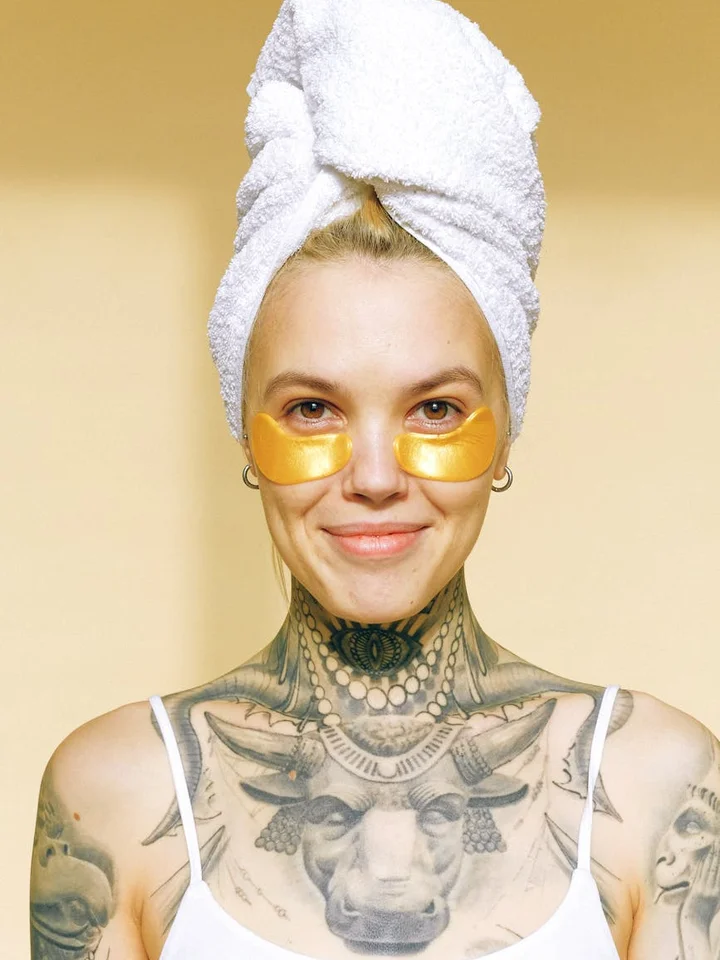 Person with a towel wrapped around their head, wearing gold under-eye patches, and tattooed chest and shoulders.