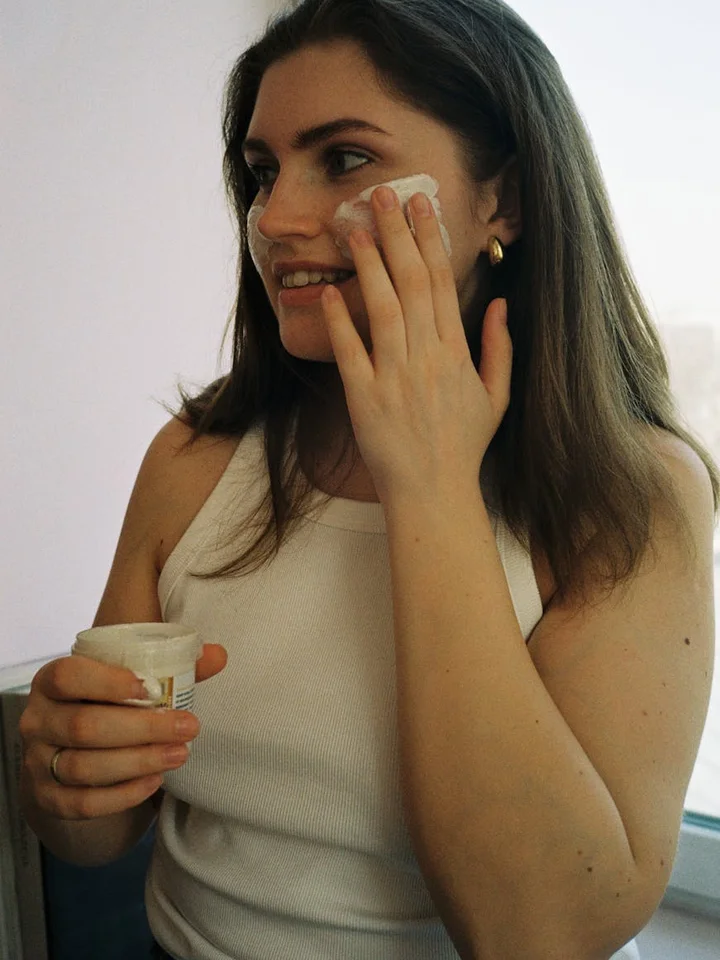 Woman applying moisturizer to her face with a small jar in hand.