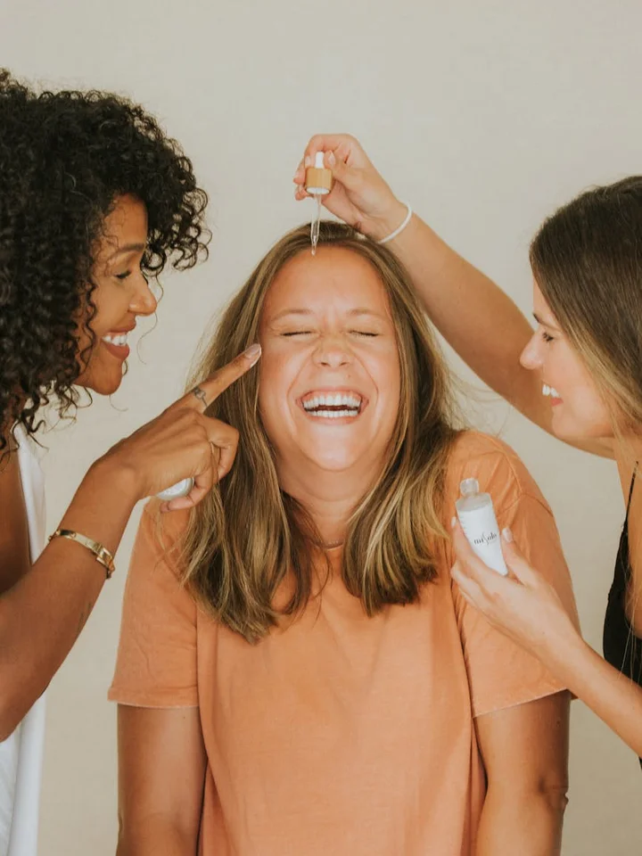 Three women in a bright room applying skincare serum to one another; a dropper is used on the central woman's forehead while two friends assist.