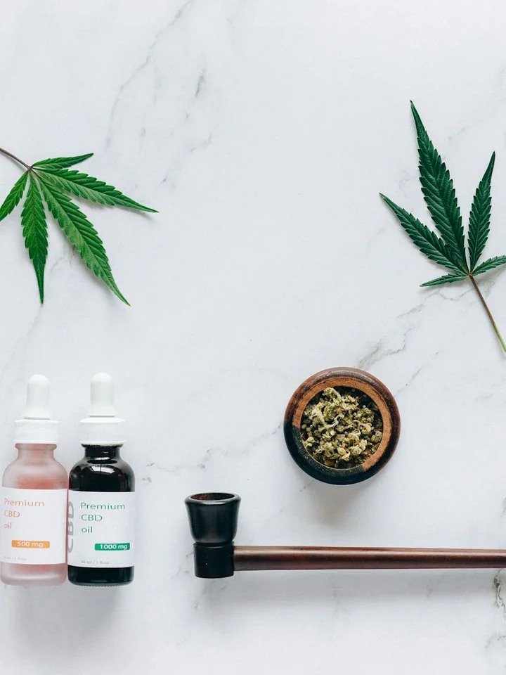 Hemp leaves, two dropper bottles of essential oil, a small bowl with hemp buds, and a glass dab tool on a white marble surface.
