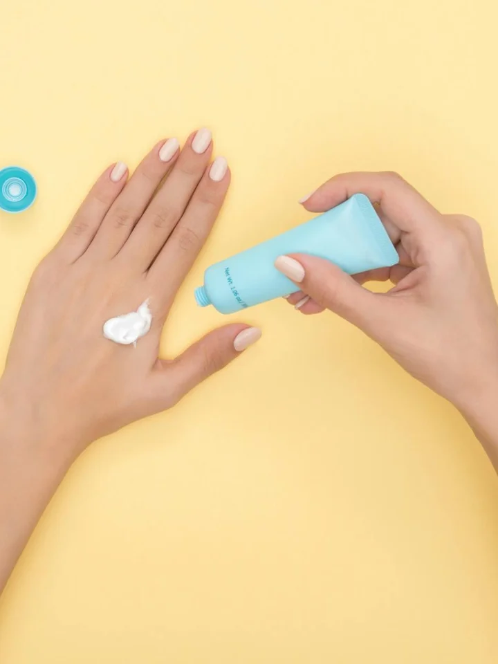 Manos aplicando crema desde un tubo azul sobre el dorso de la otra mano, con fondo amarillo