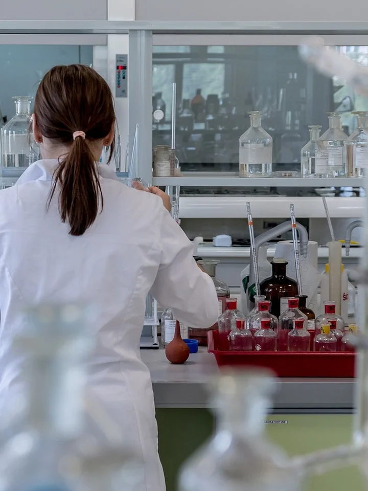 A female scientist in a white lab coat works at a laboratory bench with glassware and bottles in the background.