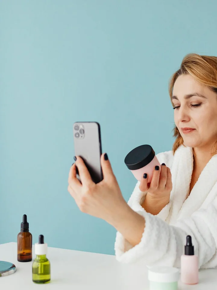 Woman in a white robe taking a selfie with skincare products arranged on a white table in front of a light blue wall.
