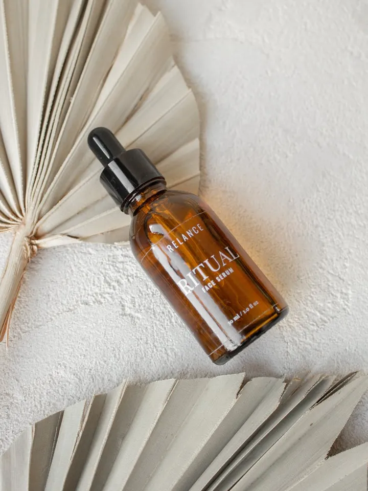 Brown glass serum bottle with a dropper on a light textured surface, with dried palm leaves in the background.