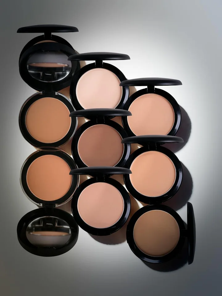 An arrangement of circular Maybelline makeup compacts with black lids placed on a reflective surface.