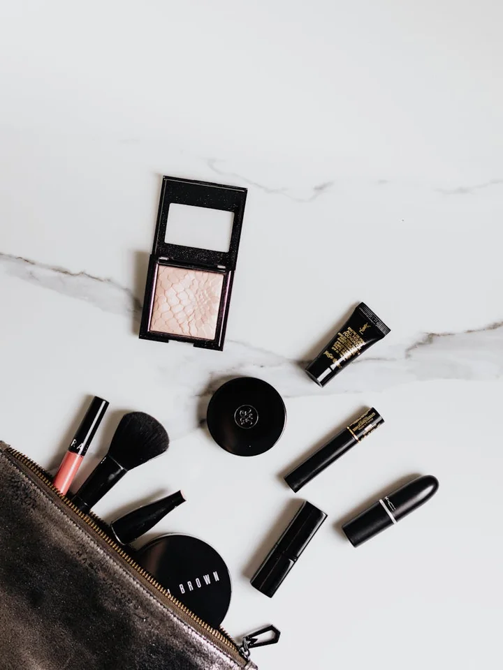 Assorted makeup items laid out on a white marble surface, including a compact, lipstick, brushes, and a small bag.