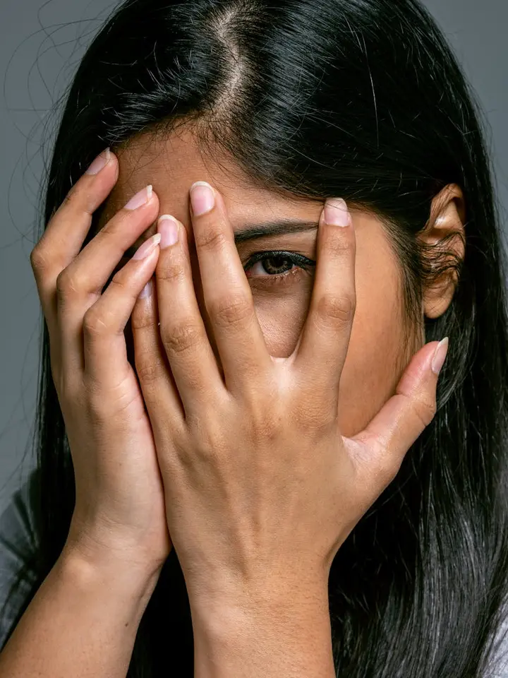 Close-up of a person with dark hair covering their face with both hands, eyes visible through the gaps between fingers.