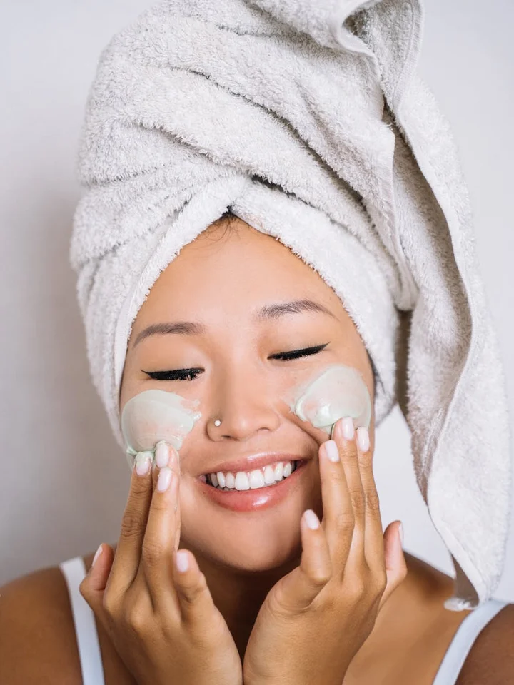 Smiling woman with a towel wrapped around her hair, applying skincare product to her cheeks.