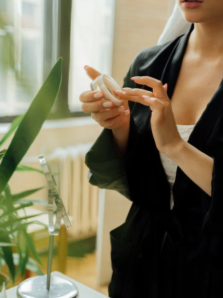 Person in a dark blazer holding an open jar of moisturizer near a mirror and plant, about to apply skincare.
