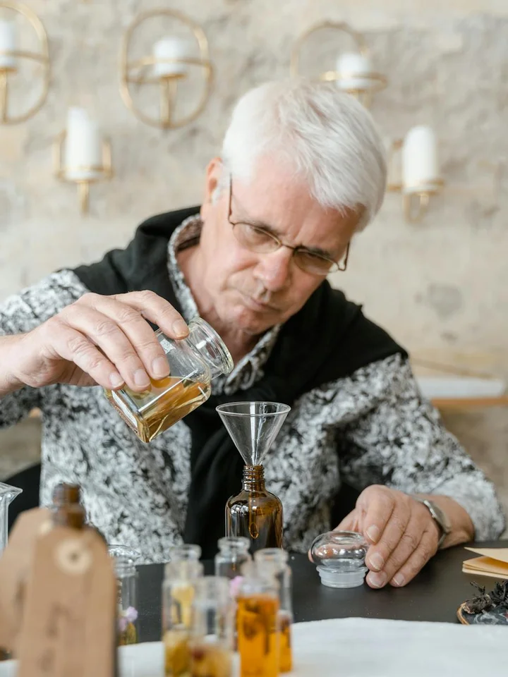 Older man with white hair and glasses pours oil from a small bottle into a glass container, surrounded by various oil bottles on a dark table in a bright, rustic setting.