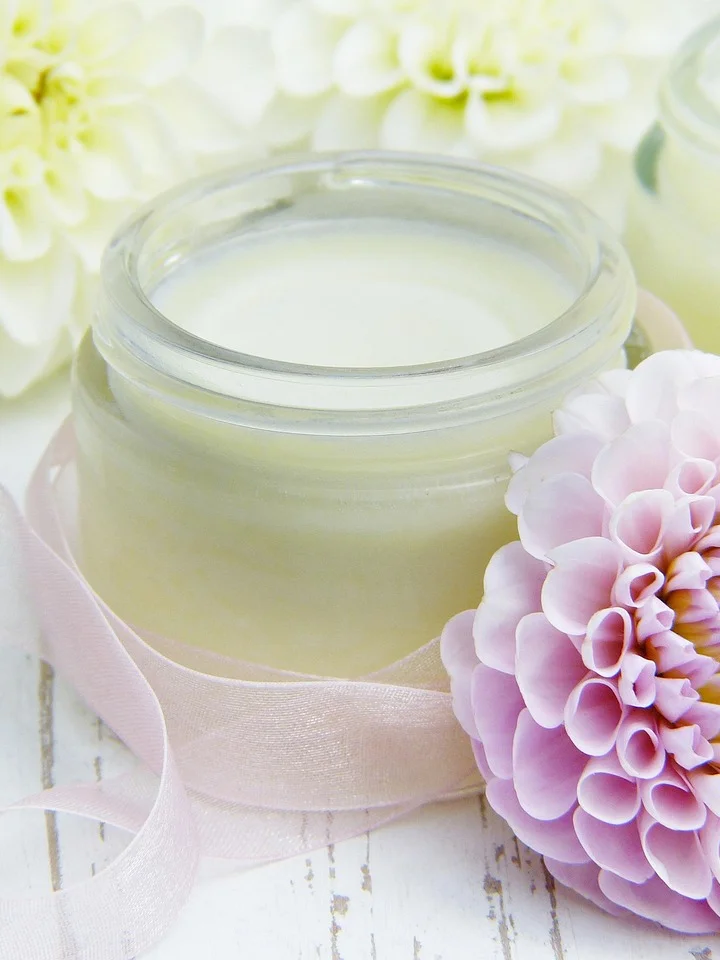 Glass jar of pale moisturizer surrounded by pink and white flowers on a light wooden surface