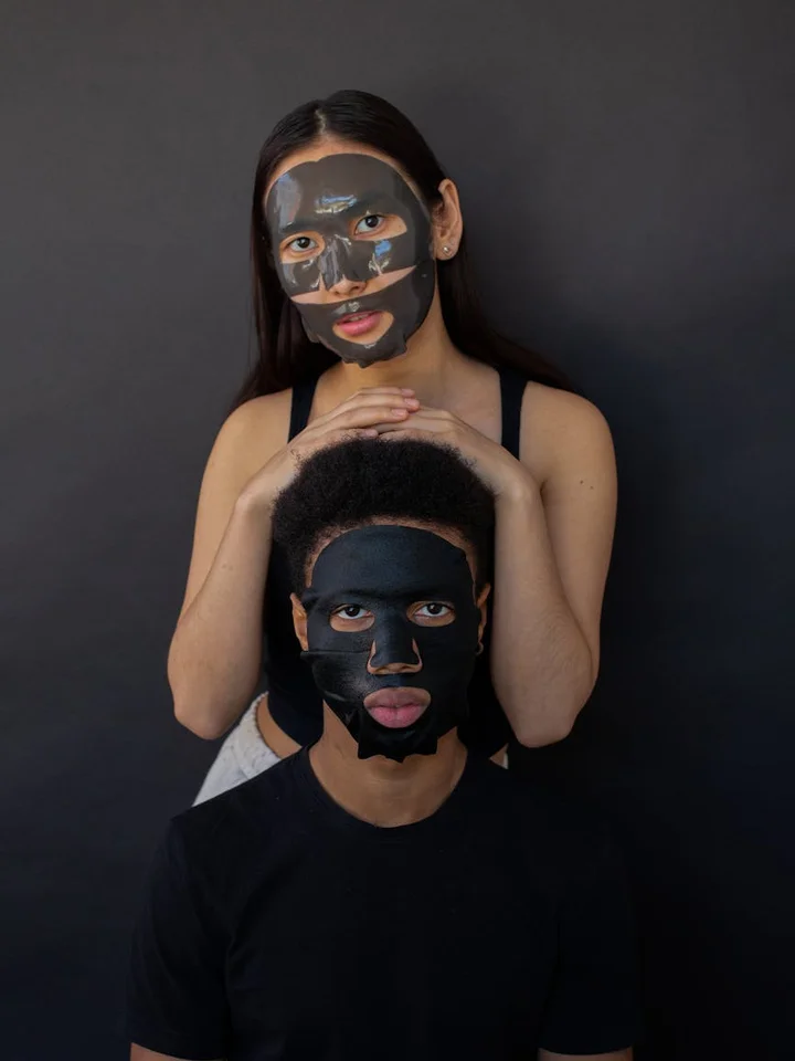 Two models wearing black peel-off facial masks pose against a dark backdrop; a person sits in front while another stands behind with their hands resting on the front person’s head.