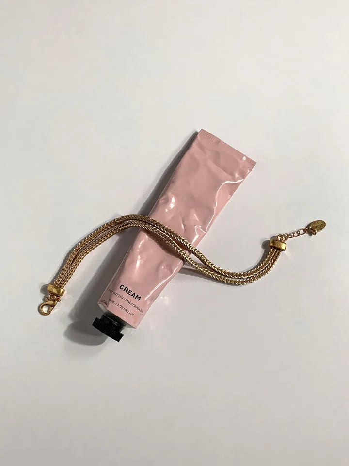 A pink tube labeled 'CREAM' sits on a white surface with a gold chain draped across it.