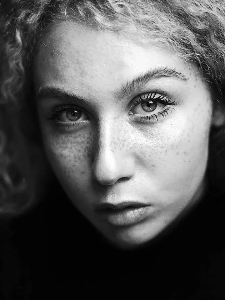 Close-up black-and-white portrait of a young woman with freckles and thoughtful expression, looking at the camera.