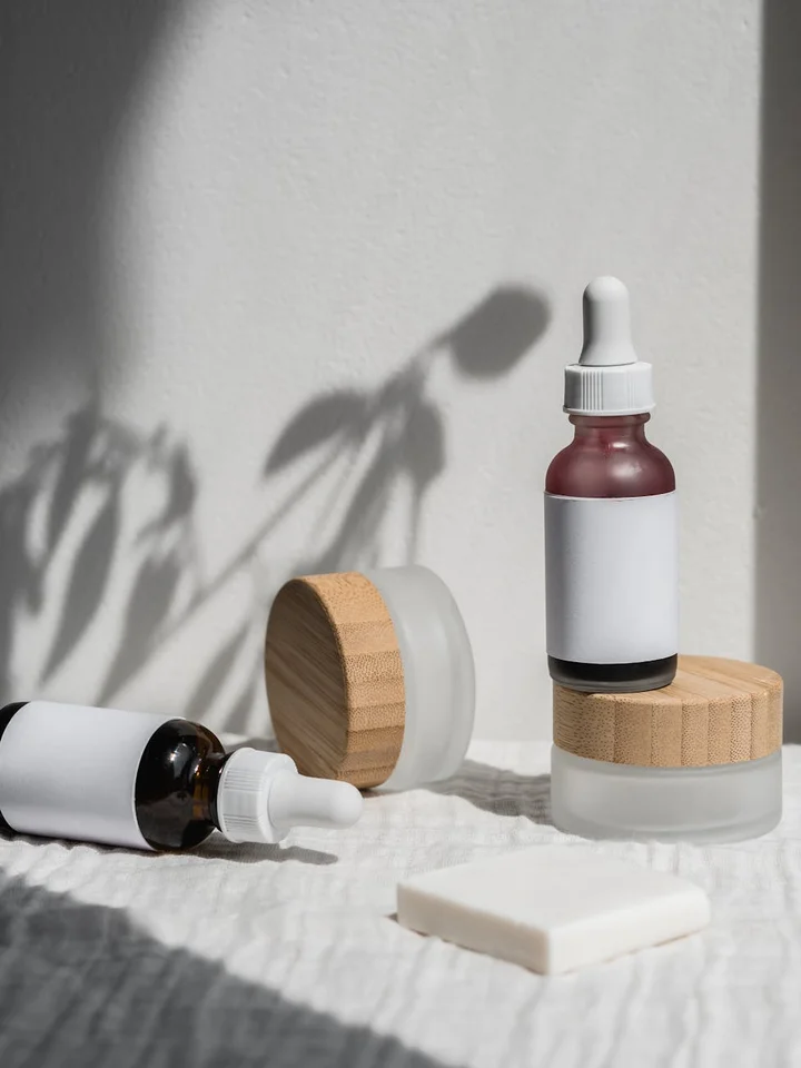 Assorted skincare containers with droppers and jars on a white textured surface; a small dropper bottle with a red tint stands upright among other containers.