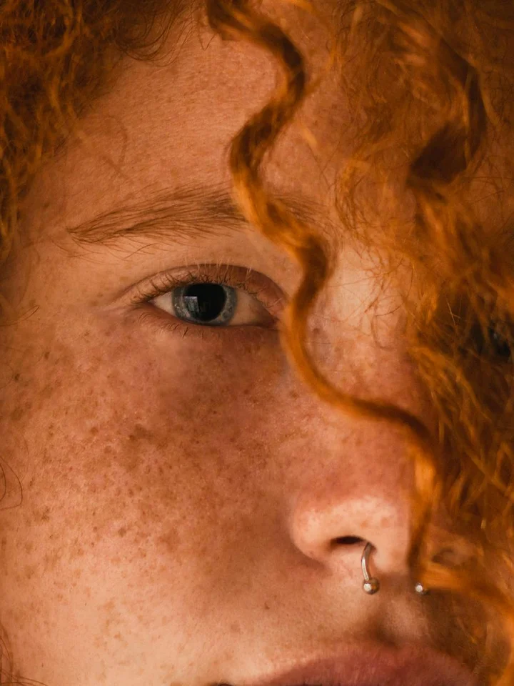 Close-up portrait of a freckled person with curly red hair and a nose piercing.