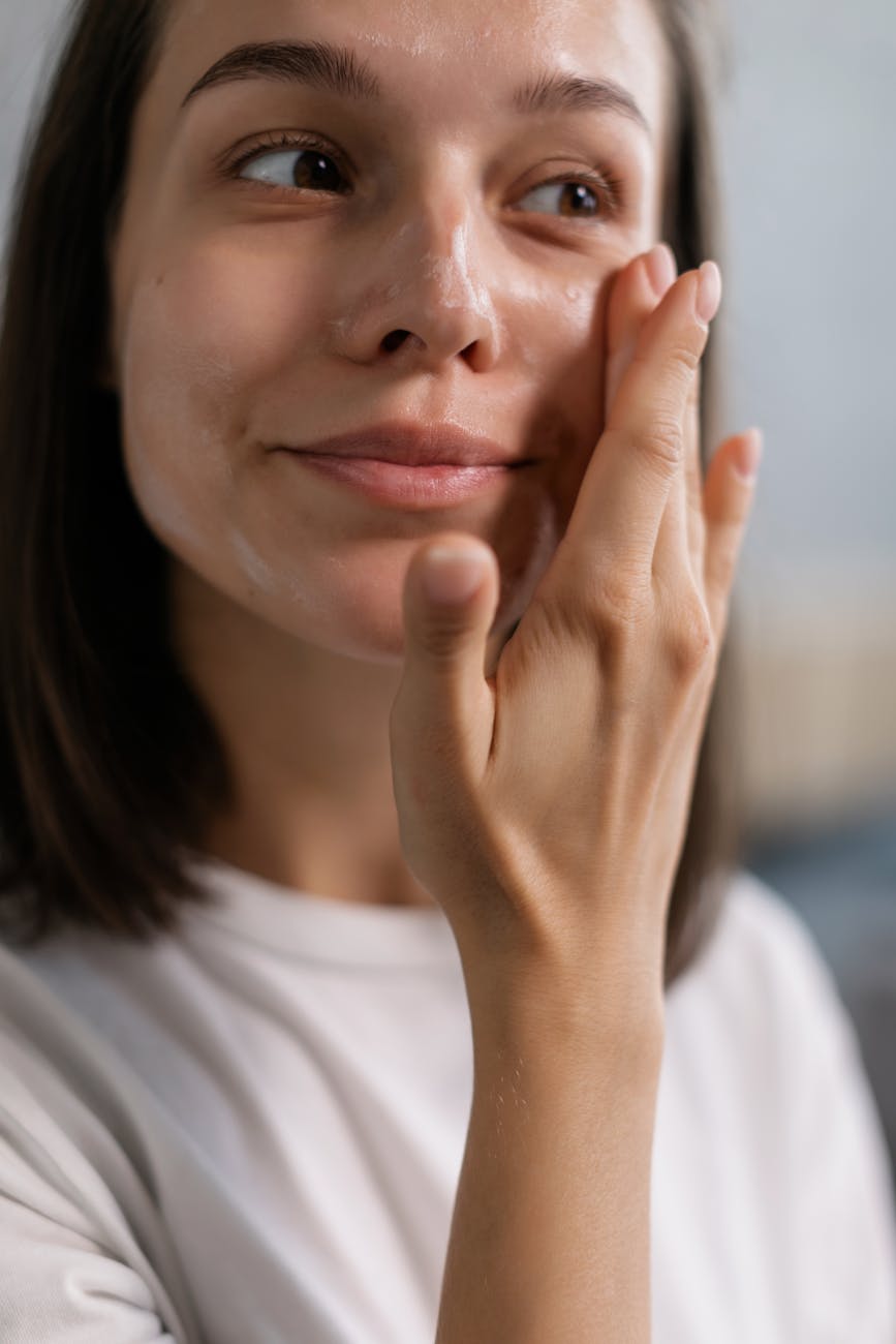Smiling woman with a towel wrapped around her head, applying foamy face wash in a bathroom.