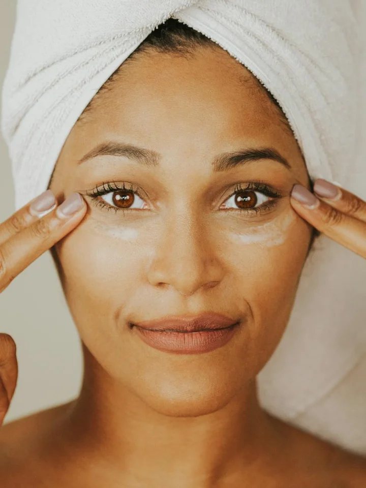 Close-up of a woman with a towel wrapped around her head gently applying skincare near her eye area.